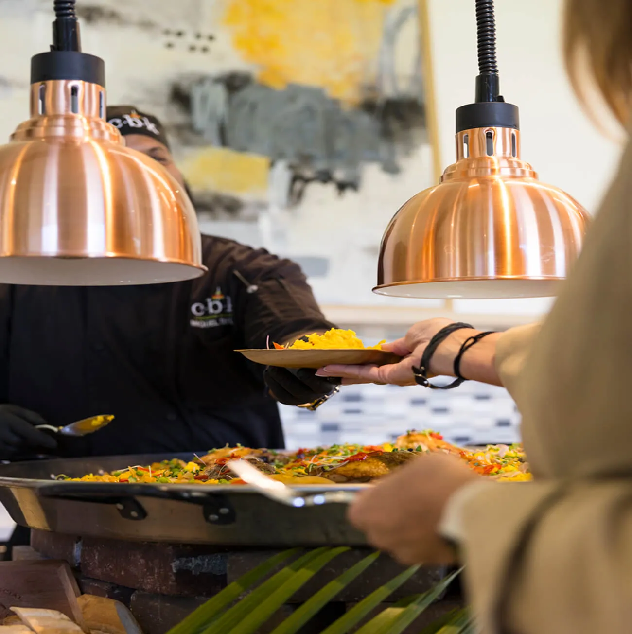 Chef serving yellow rice from a large paella pan under copper heat lamps